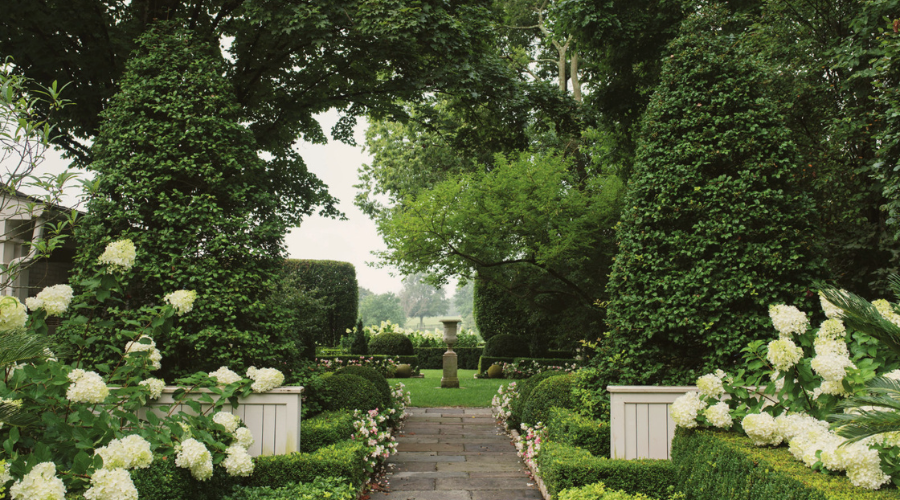 An elegantly manicured garden with a tree archway, hydrangea bushes, and a symmetrical pathway leading to a central focal point.