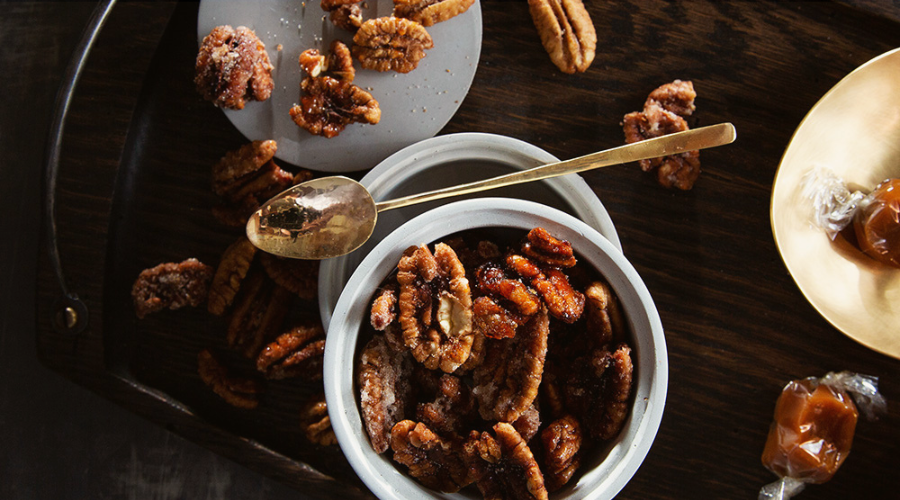 A bowl of candied pecans on a wooden surface with some nuts scattered and a spoon resting beside it.