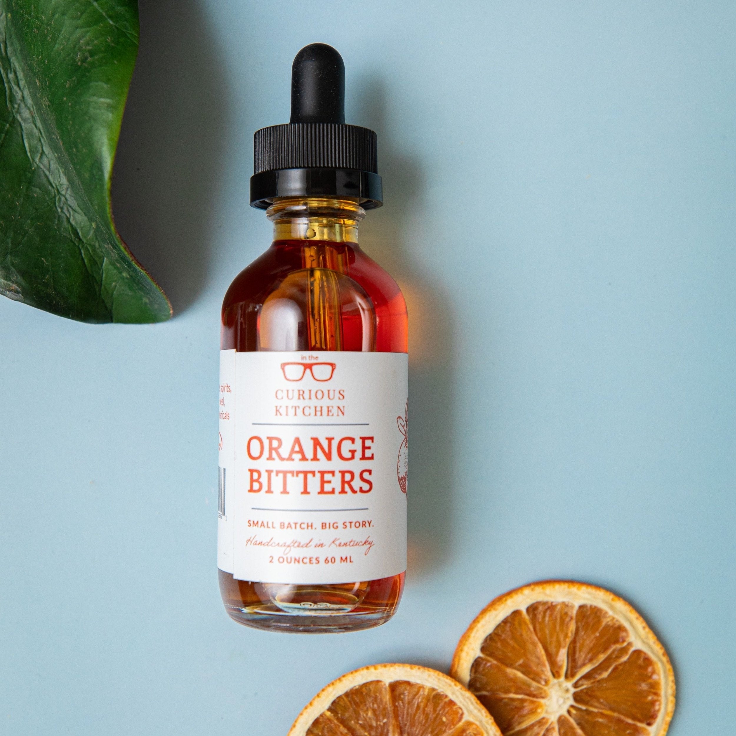 Bottle of Curious Kitchen Orange Bitters with oranges on a light blue background