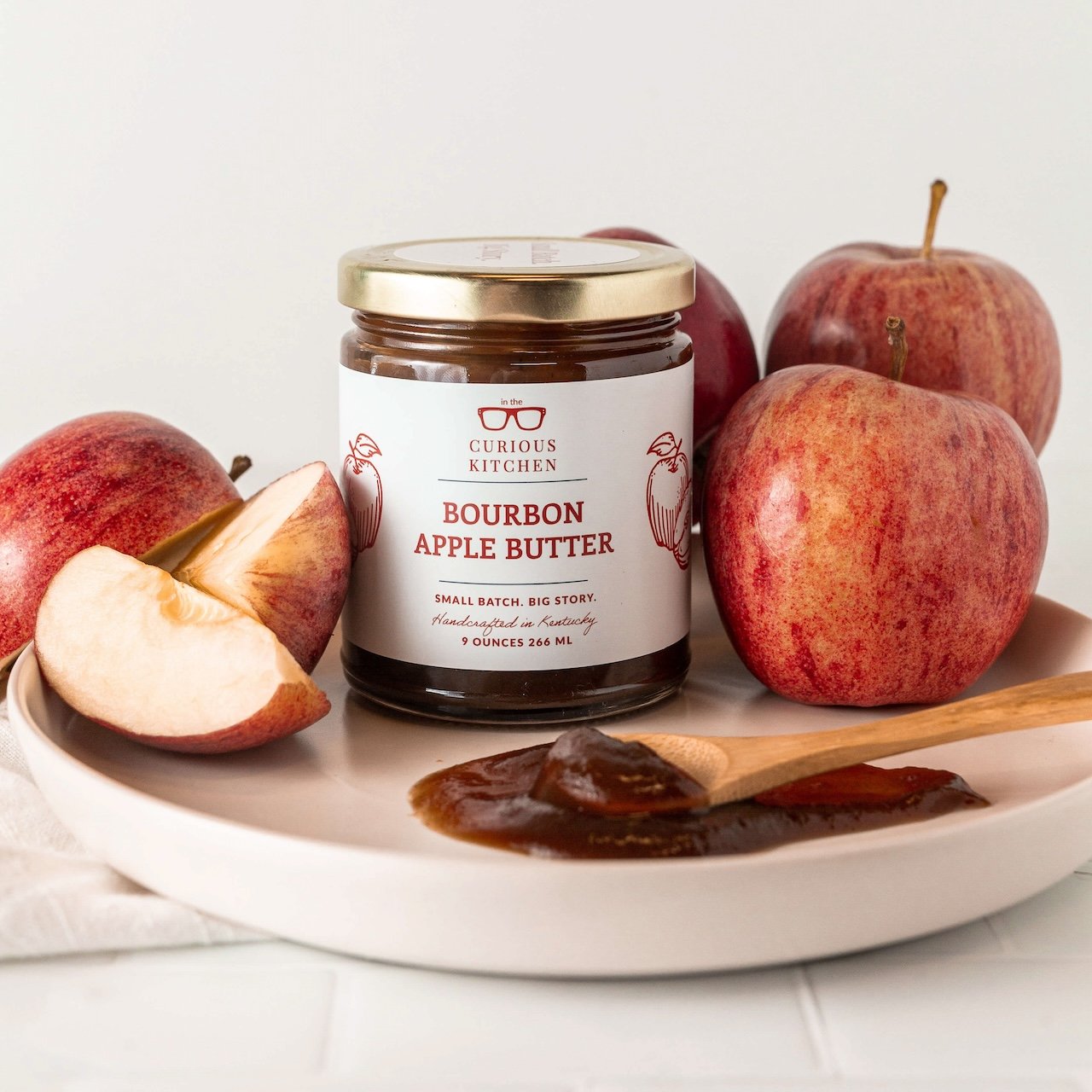 Jar of Curious Kitchen Bourbon Apple Butter with apples and a wooden spoon on a plate.