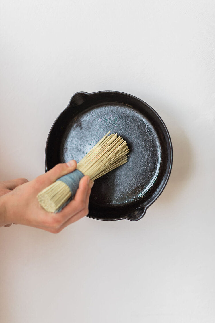 Person using a brush to apply a substance to a black cast iron skillet on a white background