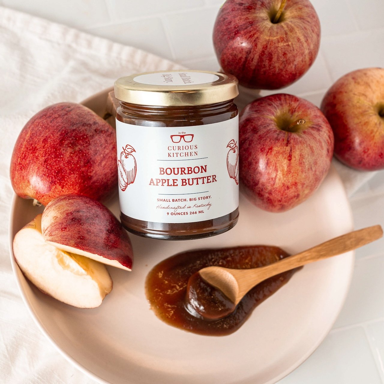 Jar of Bourbon Apple Butter from Curious Kitchen surrounded by apples on a plate.