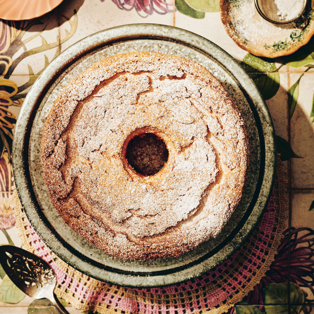 A freshly baked bundt cake with a dusting of powdered sugar on a plate, surrounded by rustic kitchen decor and warm sunlight.