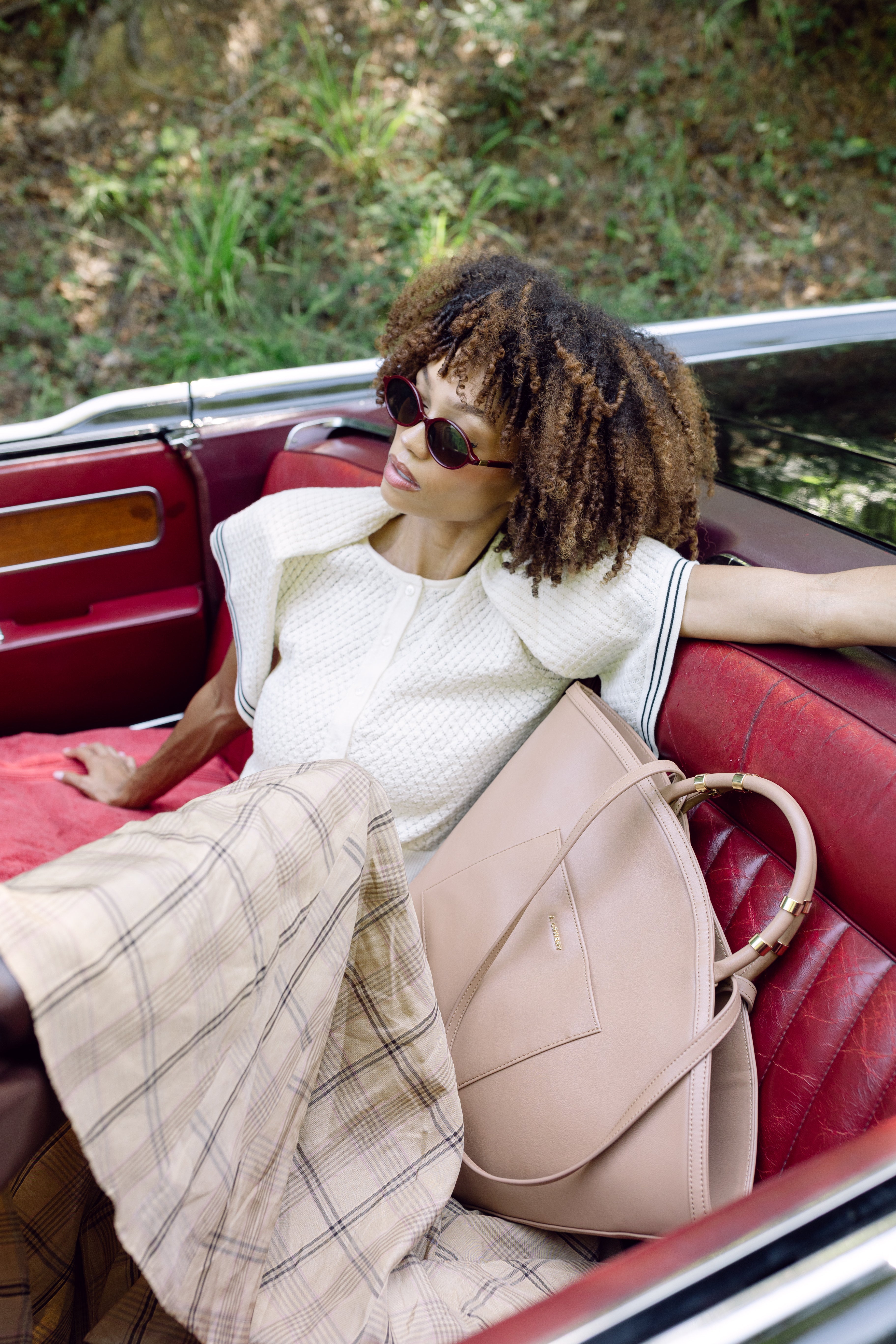 Woman sitting in a vintage car with a beige backpack, wearing sunglasses and a plaid skirt.