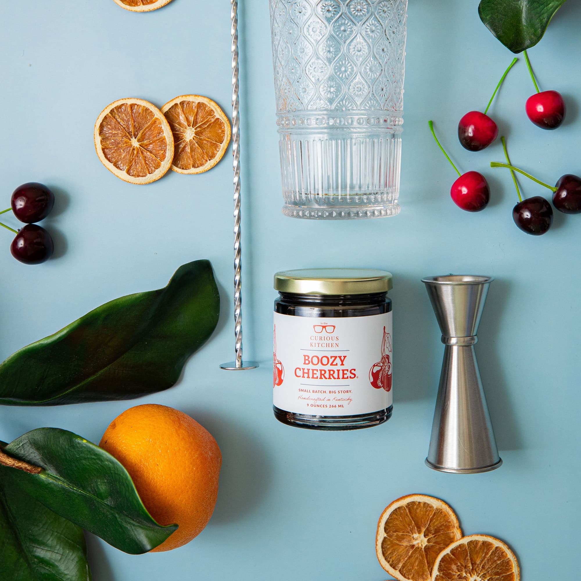 Jar of 'Boozy Cherries' with fruit and bar tools on a blue background
