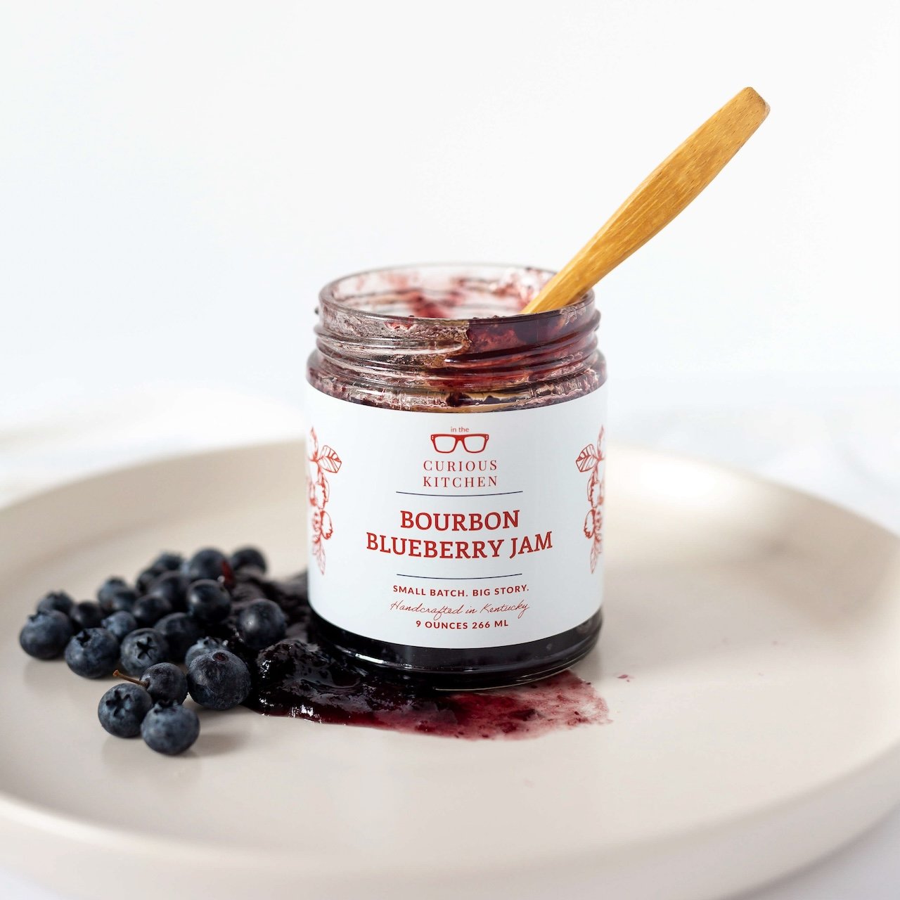 Jar of Bourbon Blueberry Jam with a wooden spoon on a white plate with blueberries.