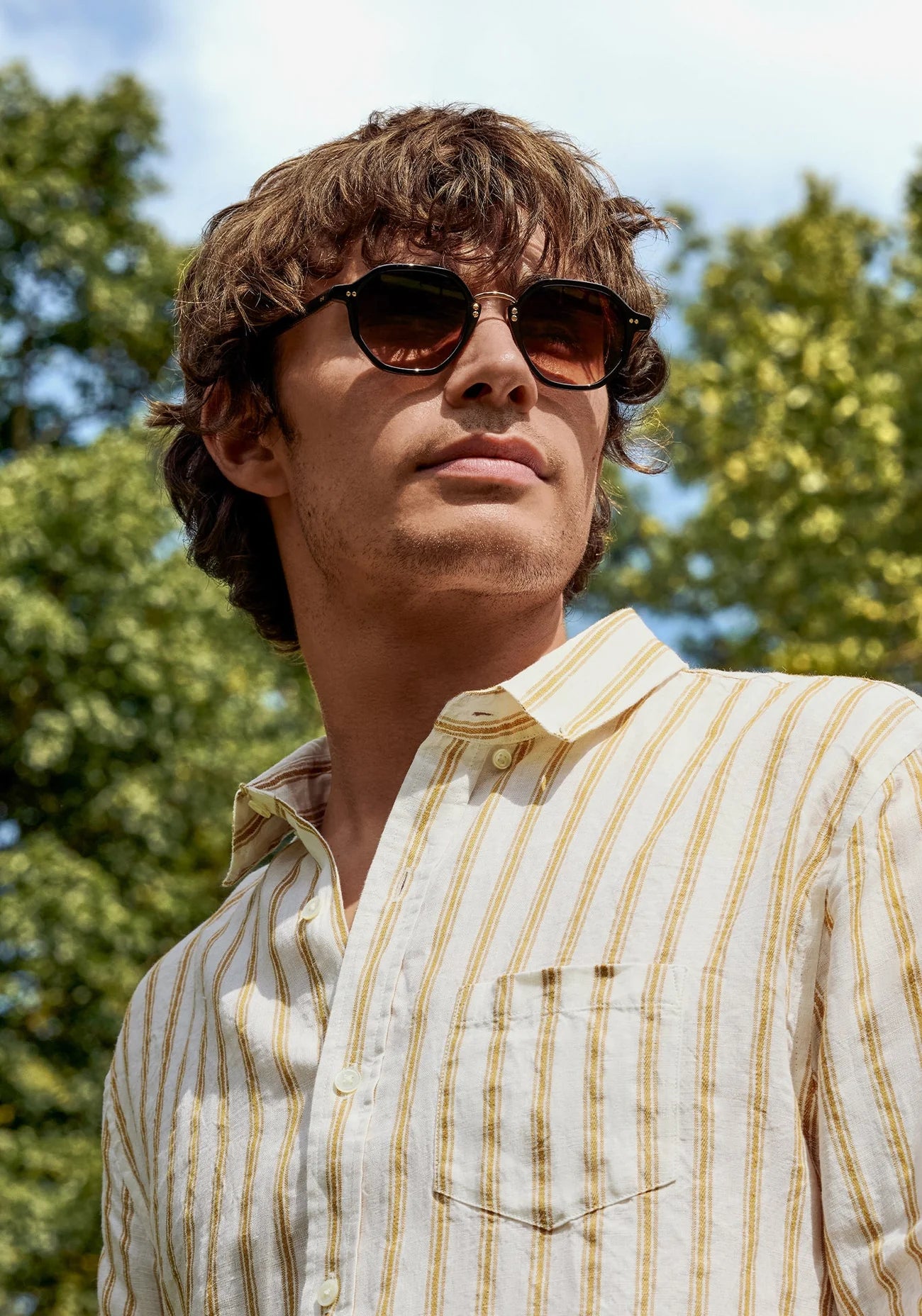 Man wearing sunglasses and a striped shirt outdoors with trees in the background