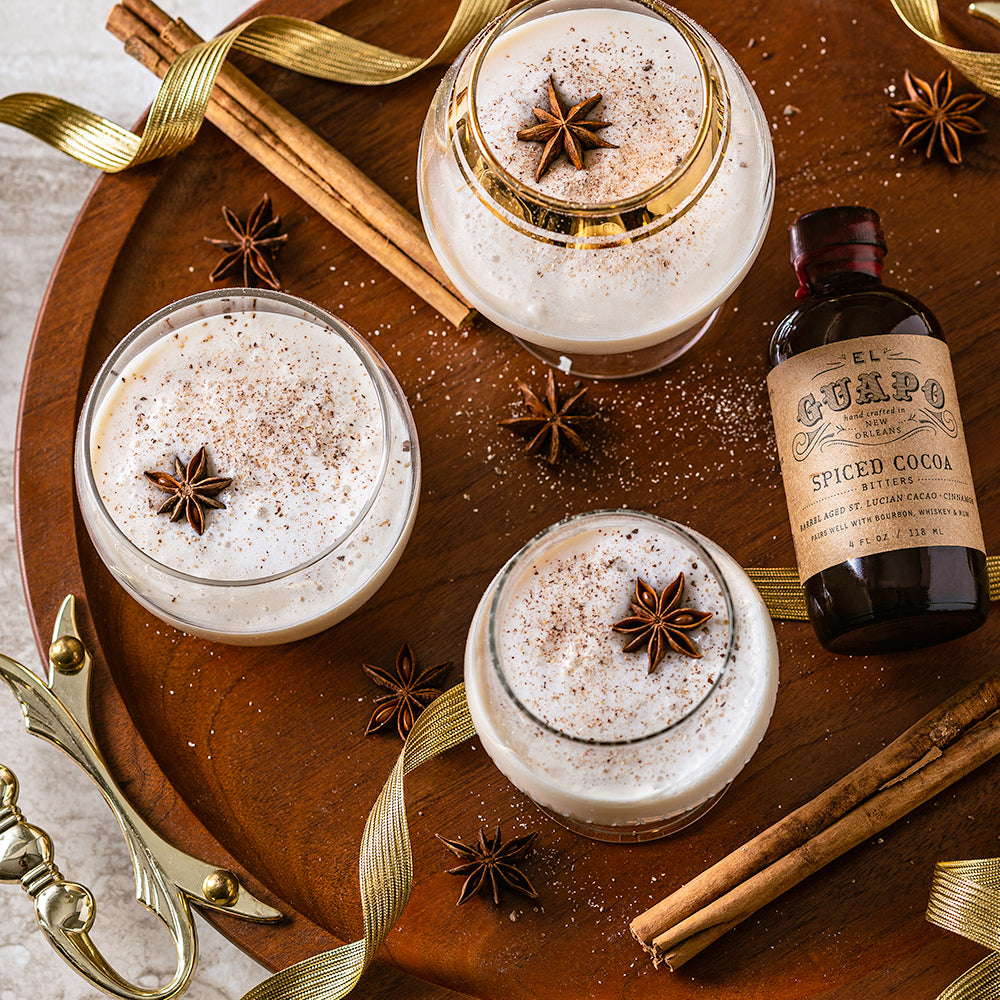A bottle of Spiced Cocoa Bitters placed next to glasses filled with a cocktail garnished with cinnamon sticks and star anise on a wooden surface.