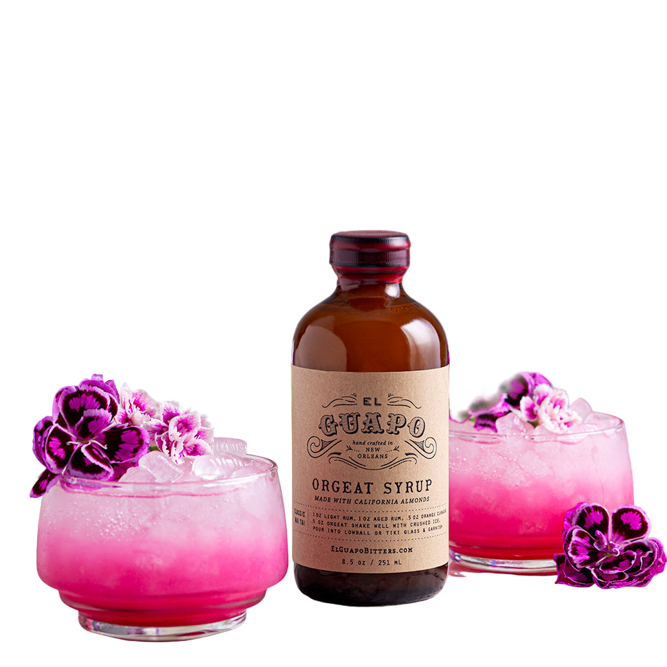 A bottle of Orgeat Syrup with two glasses of a pink cocktail garnished with purple flowers in the background, labeled 'EL GUAPO'.