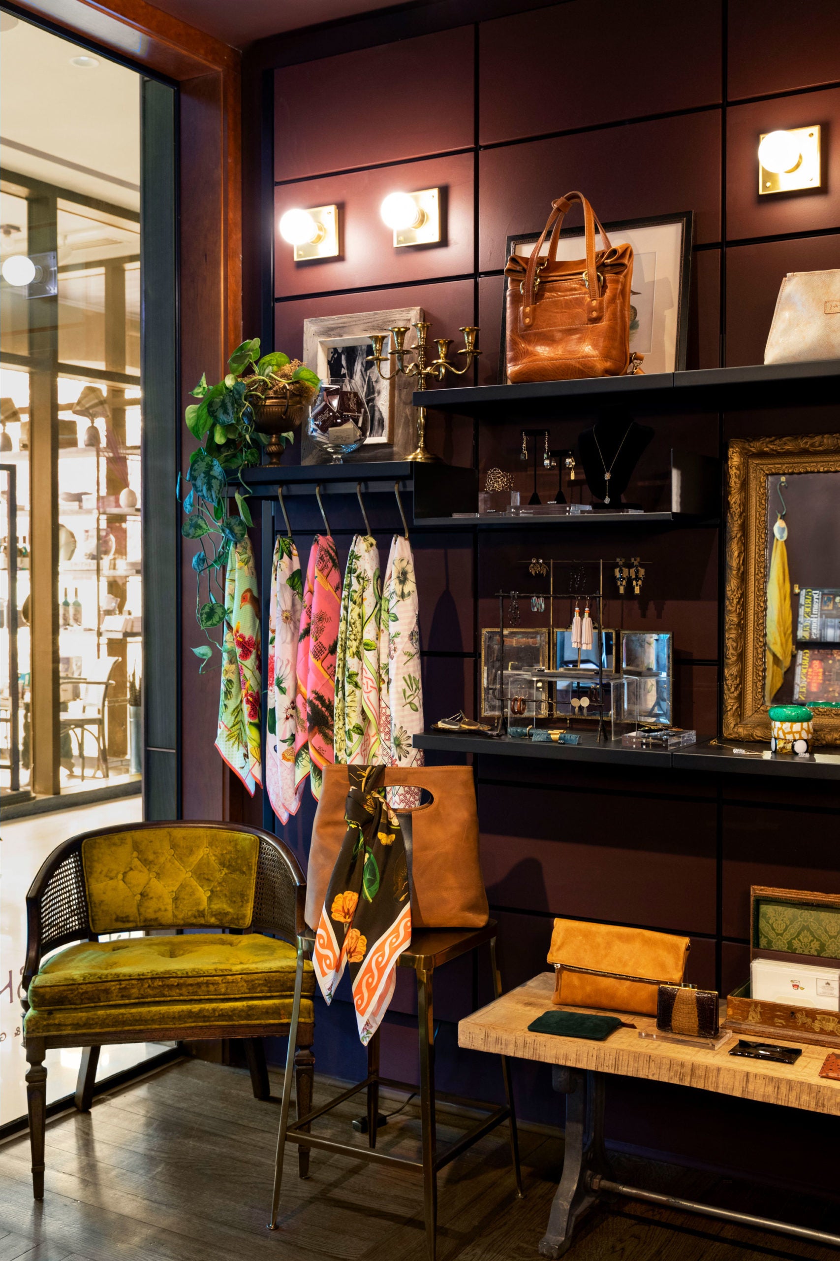 An elegant boutique interior with a vintage aesthetic, featuring a display shelf with bags and accessories, framed artworks, and a cozy seating area with a plush green chair.