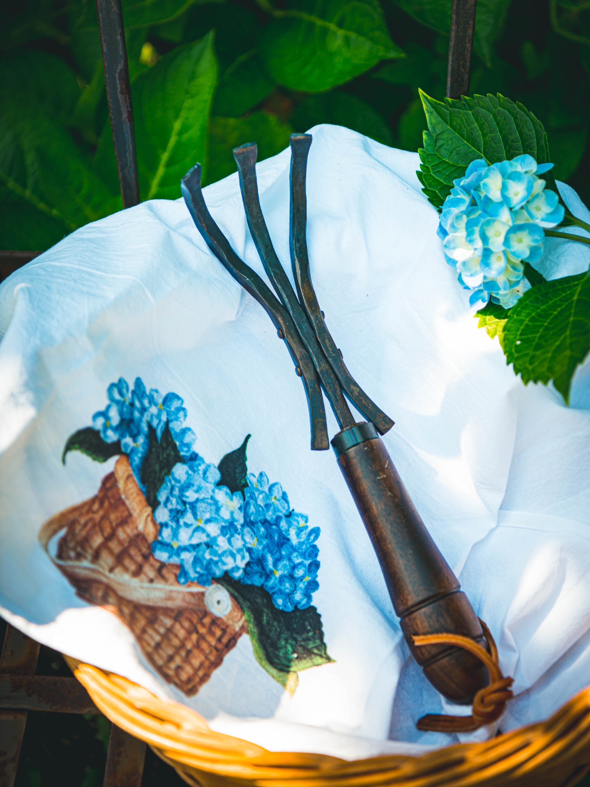 Rake and garden tools on a white cloth with blue hydrangea design, surrounded by greenery.