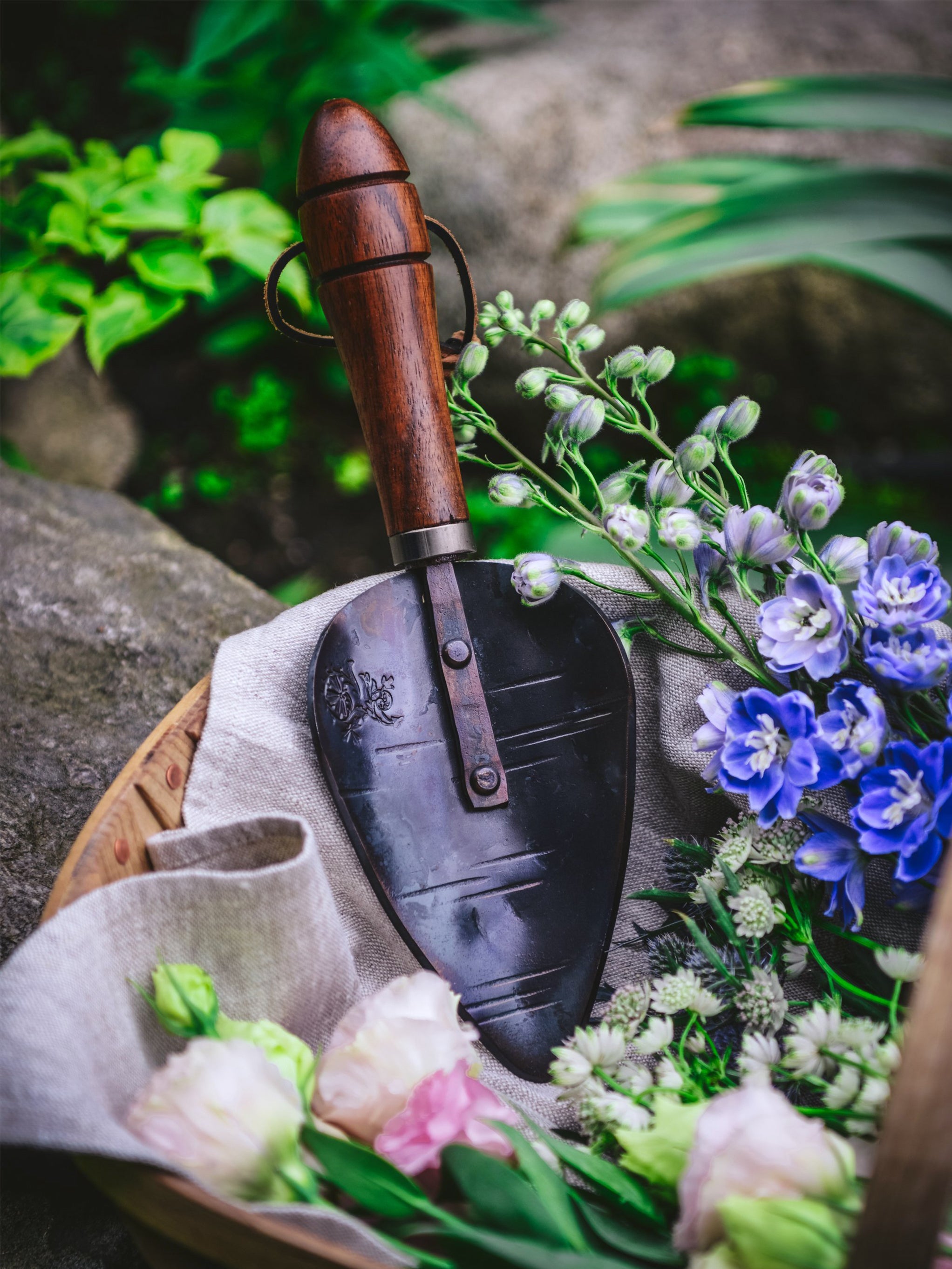Gardening trowel with wooden handle on a stone surface with flowers and plants.