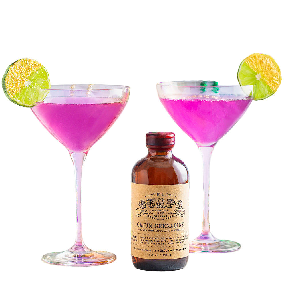 Two martini glasses filled with pink Cajun Grenadine cocktail, garnished with lime slices, with a bottle of Cajun Grenadine syrup in front, against a background of colorful beads and a marble pattern.