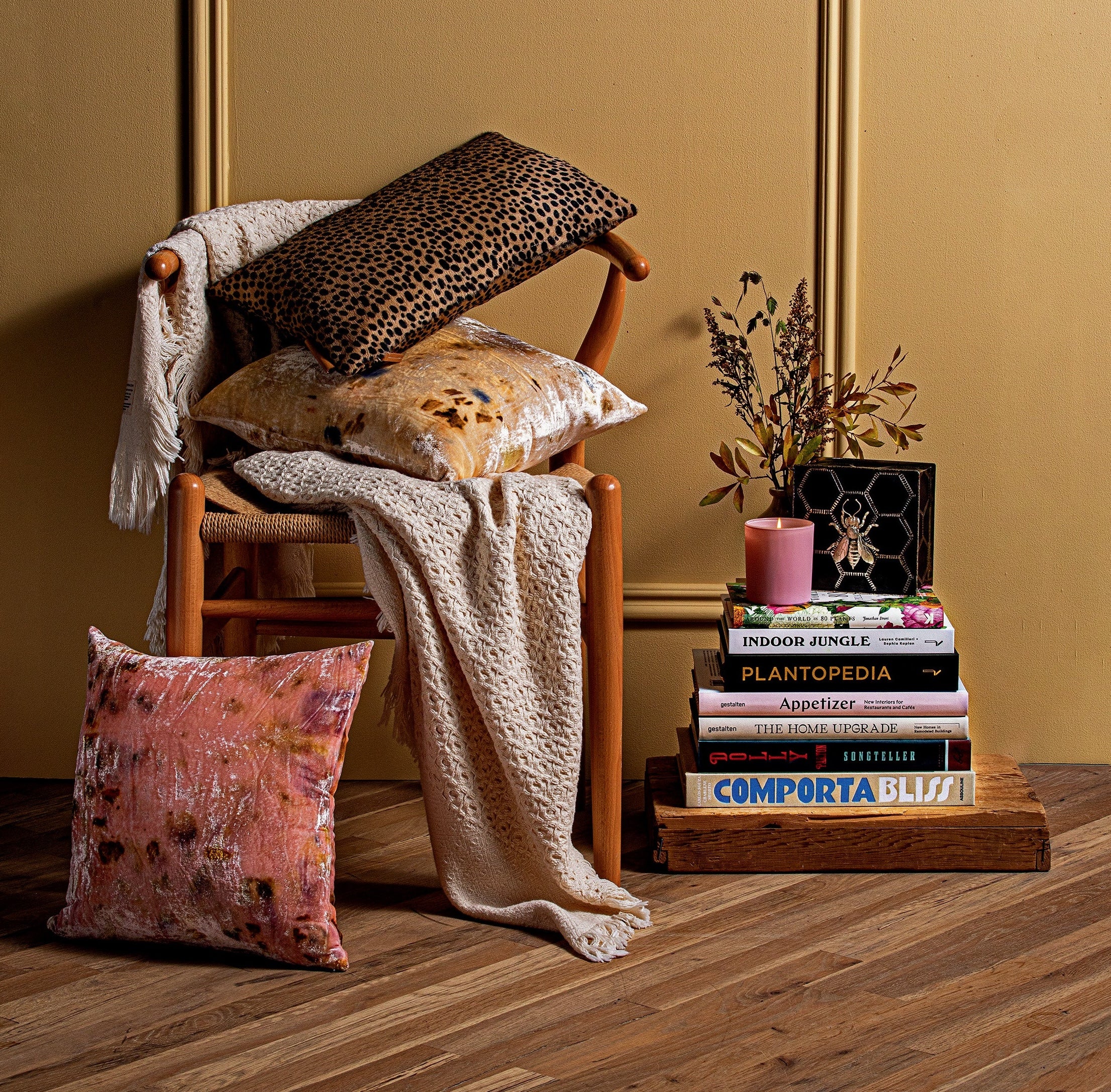 A cozy reading nook with a wooden chair adorned with a soft throw blanket and a leopard print cushion. A stack of books is placed next to the chair with a plant.