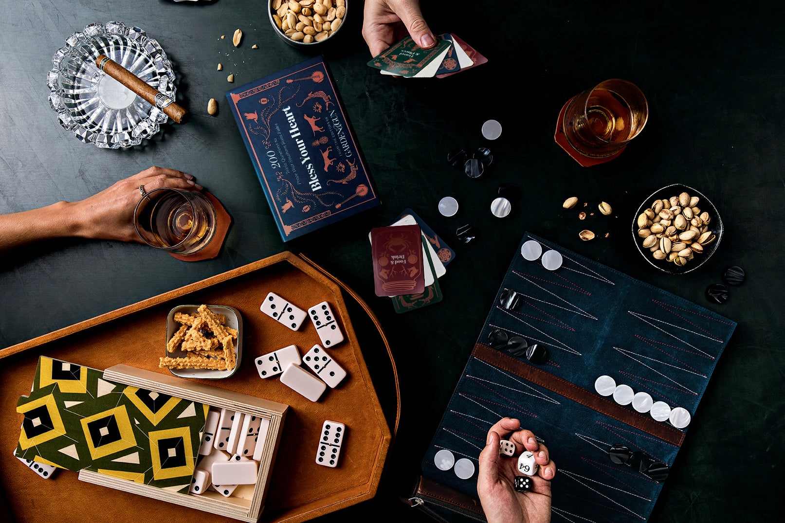A top-down view of a social gathering with hands playing backgammon, with snacks, drinks, and playing cards on the table.