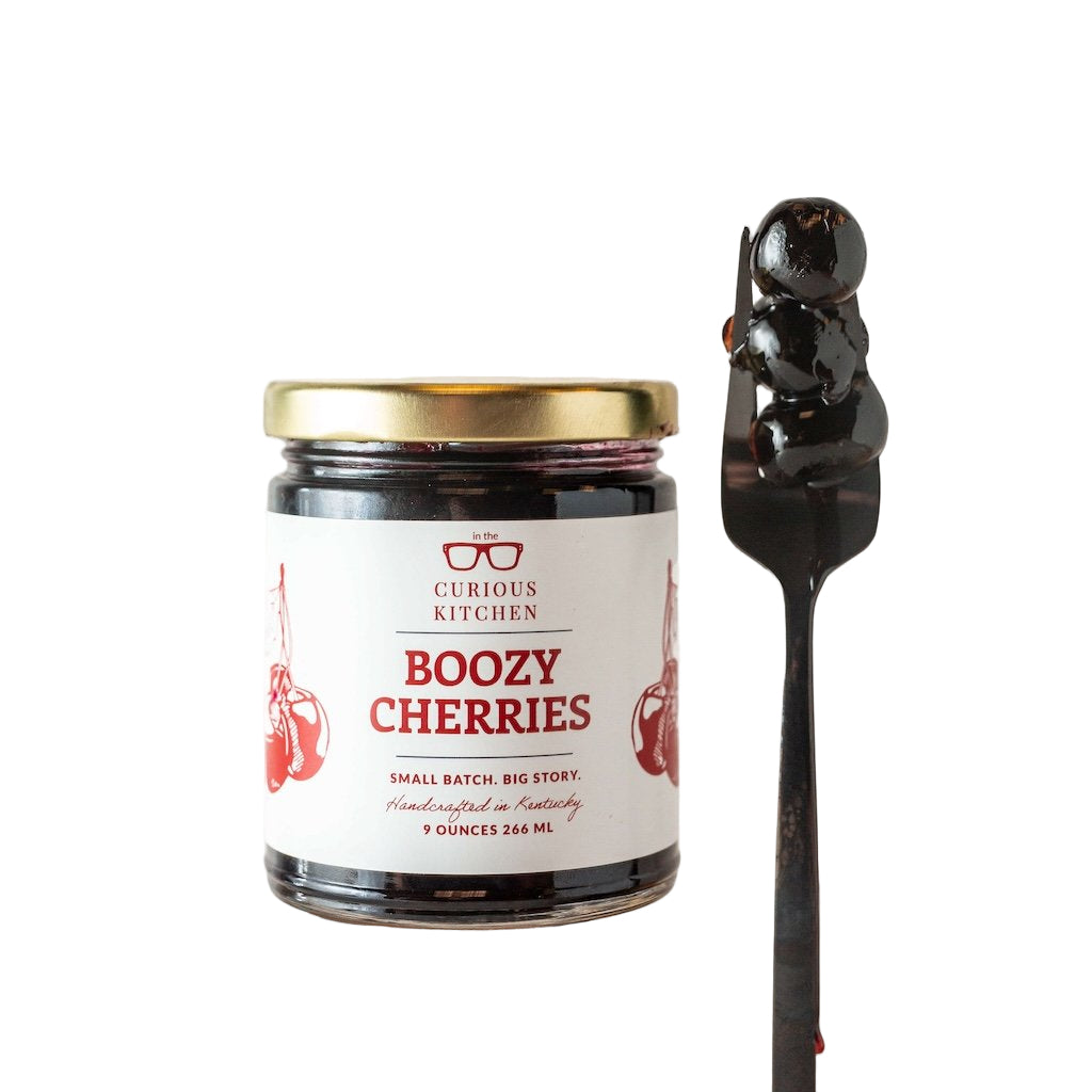 Jar of 'Boozy Cherries' with a spoon filled with cherry mixture on a white background