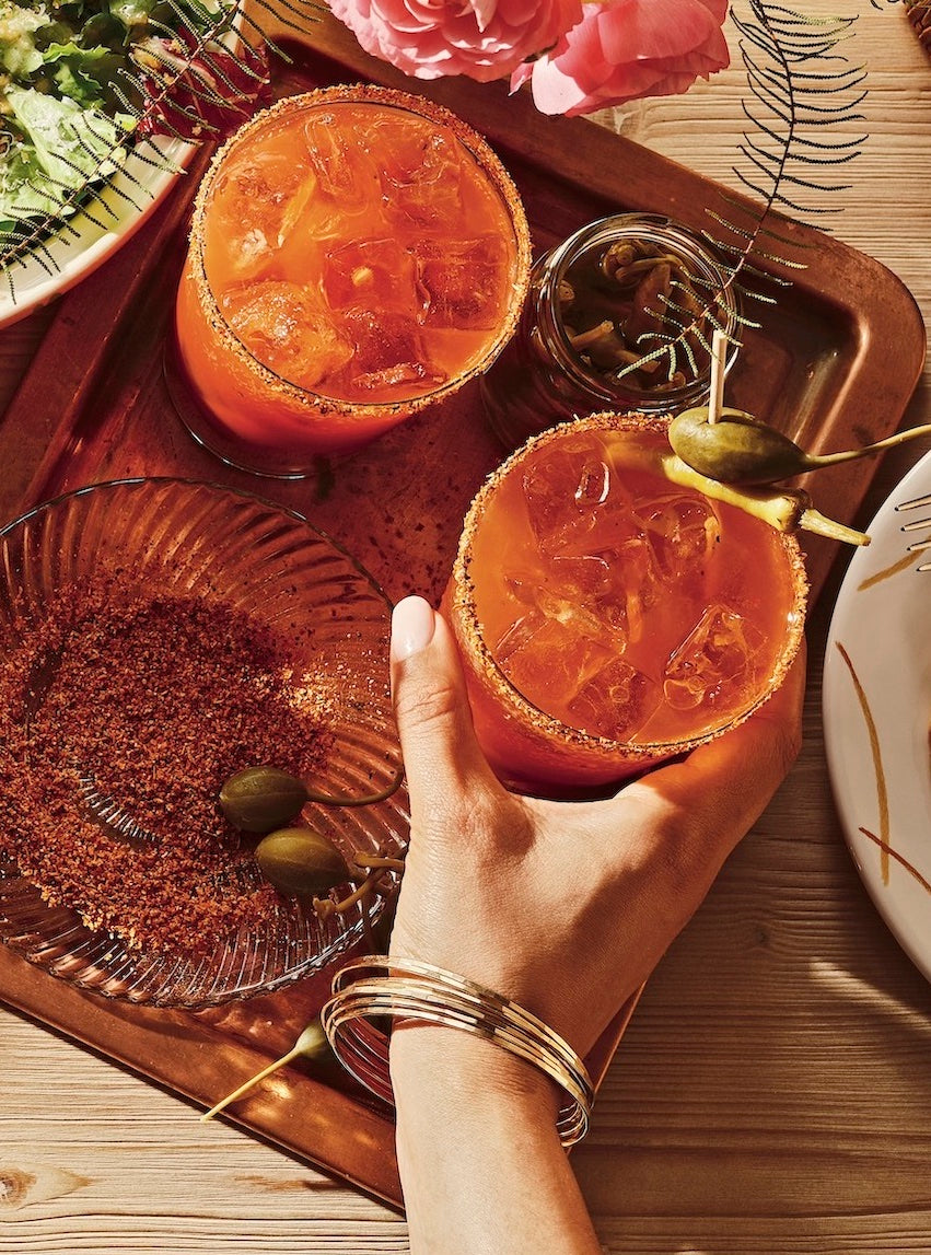 Hand holding a bloody mary glass garnished with olives, drinks, snacks, and flowers on a wooden table.